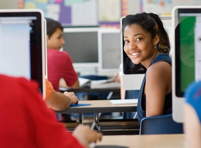 Students at desks using desktop computers in a classroom; bulletin board in background, one student looking back smiling at the camera.