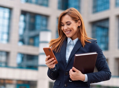 Young woman in business attire smiling while looking at her phone, holding a folder, standing outside an academic building.