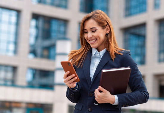 Young woman in business attire smiling while looking at her phone, holding a folder, standing outside an academic building.