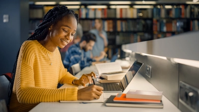 Woman studying with laptop and notebook in library.