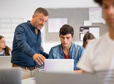 Male teacher helping student with laptop in classroom.
