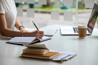 Person writing at desk with laptop and coffee.