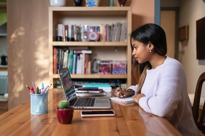 Young women working on a laptop, writing on a notebook in an office.