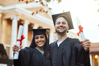 Two graduates holding diplomas in front of university building.