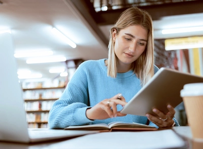 Young woman using tablet with book, laptop, and coffee in library.