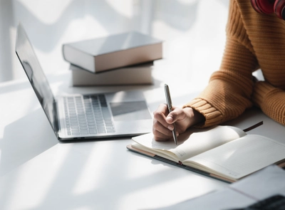Person writing in notebook at desk with laptop and books.