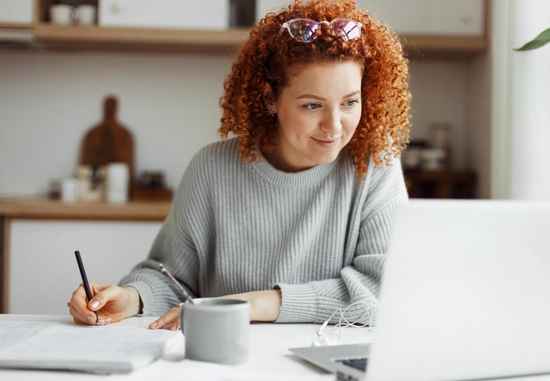 Young woman writing at table with laptop and kitchen items in background.