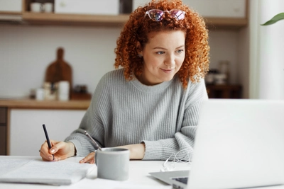 Young woman writing at table with laptop and kitchen items in background.