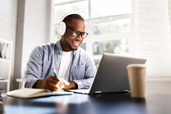 Young man with headphones writing at desk with laptop.