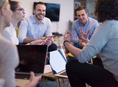 A group of people sitting in a circle having a discussion, with laptops and notebooks, and a TV screen in the background.