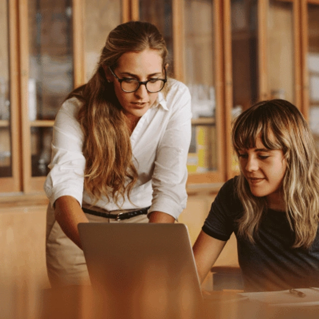 A woman helping a student with an online assignment, they are both looking at the computer screen