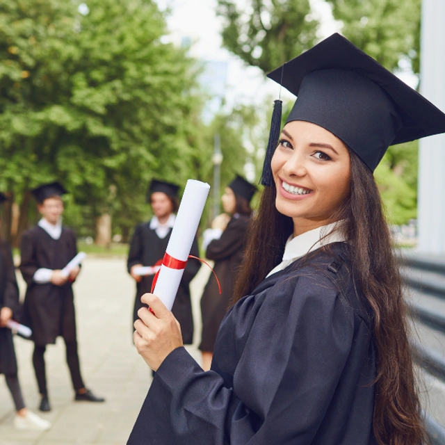 A woman with a graduation outfit looking at the camera holding a diploma, there are other graduates in the background
