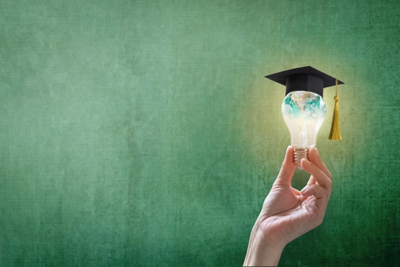 A hand holding up a lightbulb with a graduation cap on it against a green background.