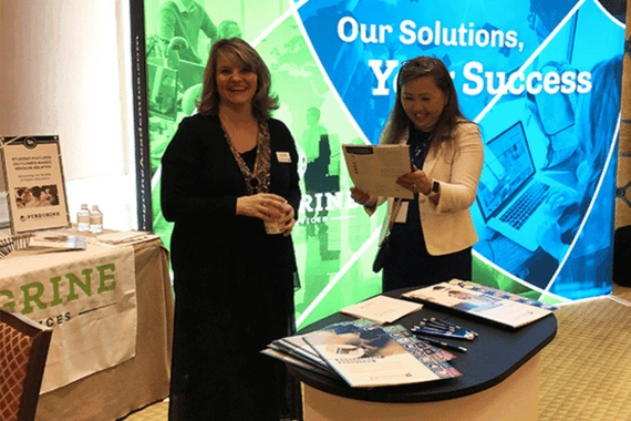 Lara Miller and Alimaa Jamiyansuren at IACBE ACAM 2019. They're standing in front of a colorful sign.