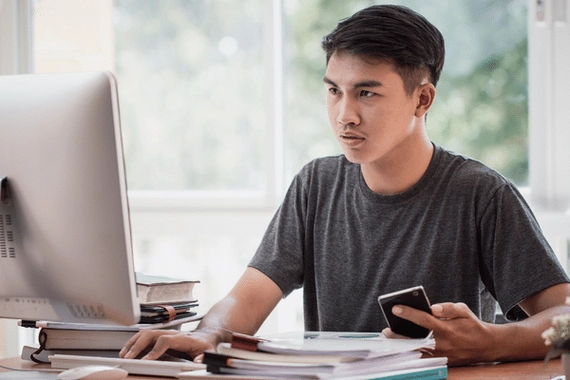 A male working at a computer. He is focused and is typing on the keyboard with one hand and holding his phone in the other.
