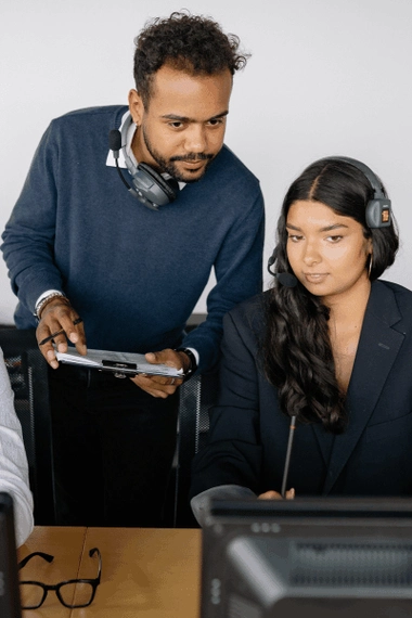 A man and a woman are looking at a computer screen. Both are wearing headphones.