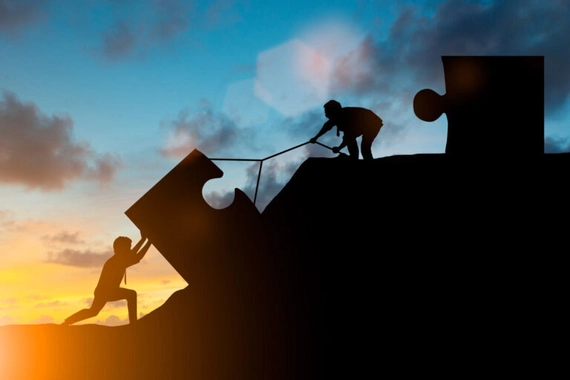 Sihouettes of two people on a hill. One person is at the top and pulling a large puzzle piece with rope while the other is at the bottom of the hill, pushing the puzzle piece. There's another puzzle piece at the top of the hill that they are trying to connect it to.